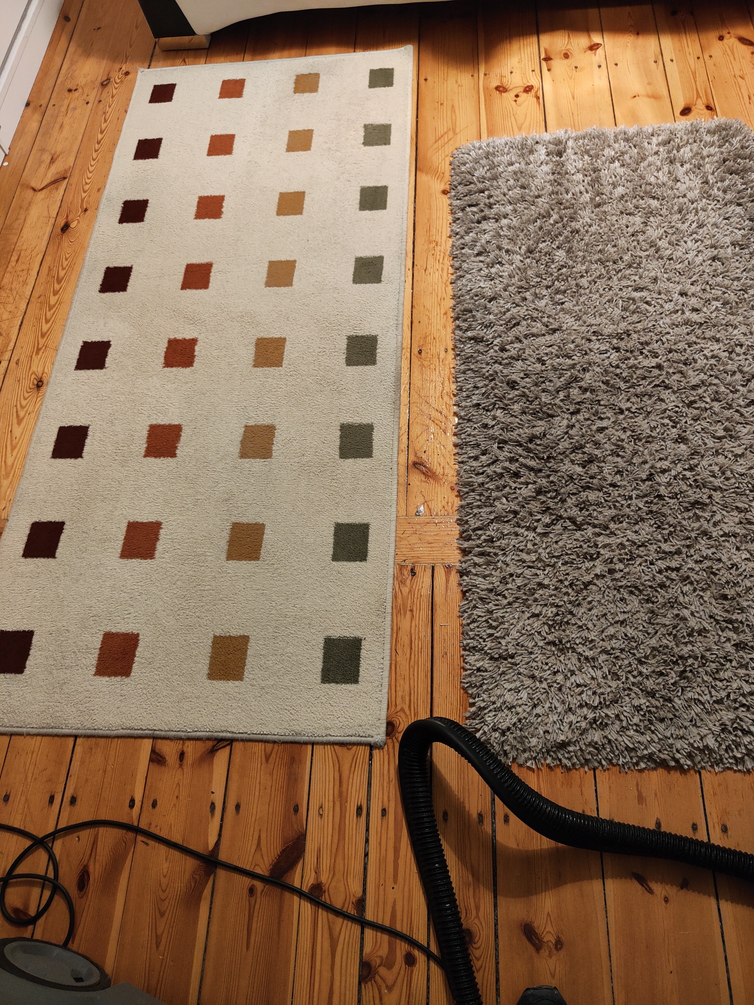 Dwa dywany na drewnianej podłodze: beżowy dywan w brązowe, pomarańczowe i zielone kwadraty oraz szary dywan typu shaggy, czyszczone odkurzaczem z widoczną rurą.