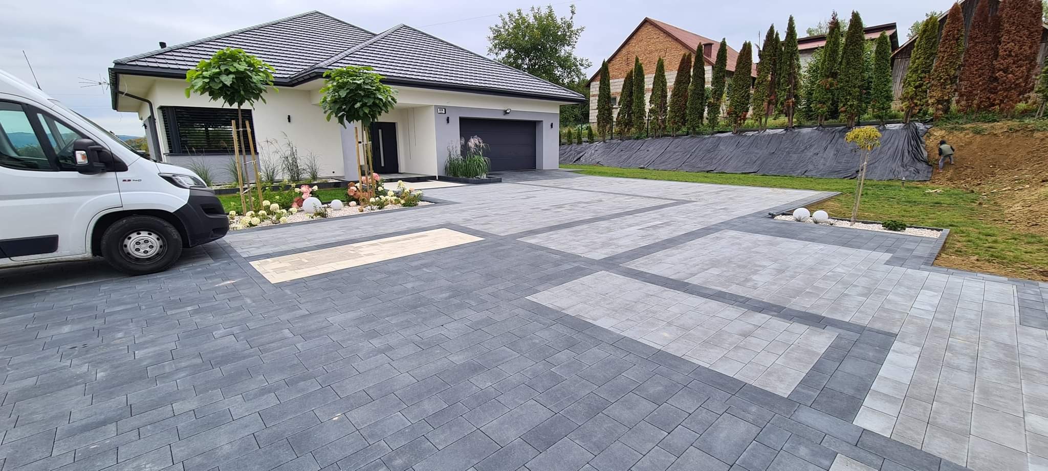 Nowoczesny podjazd z szarej kostki brukowej ułożonej we wzór prostokątów z jasnymi wstawkami, na tle domu z garażem i zadbanego ogrodu z ozdobnymi kulami.