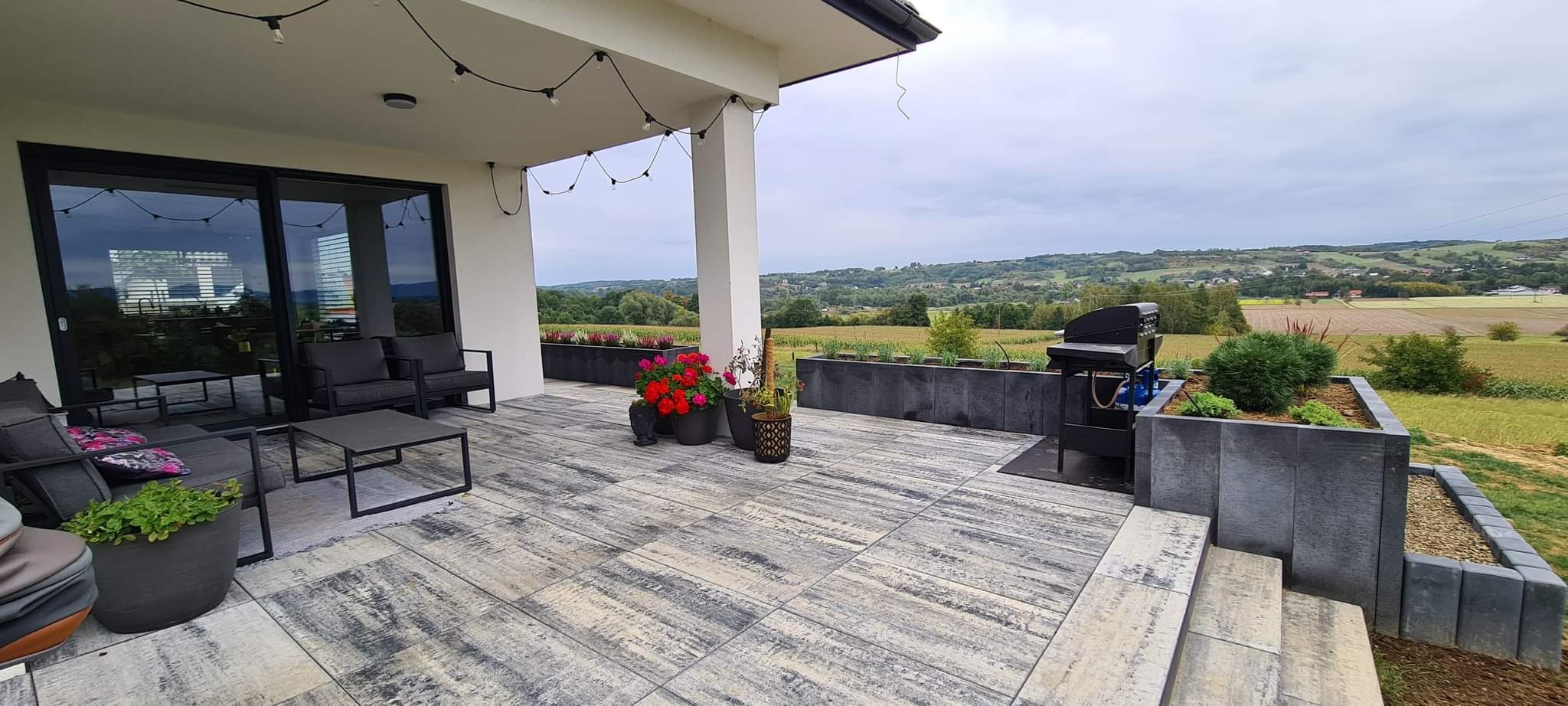 Nowoczesny taras z szaro-beżowej kostki brukowej, wyposażony w meble ogrodowe, donice z kwiatami i grill, z widokiem na krajobraz wiejski.