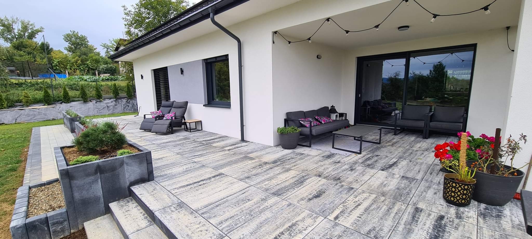 Nowoczesny taras z szarych, wzorzystych płyt betonowych, wyposażony w meble ogrodowe, rośliny w donicach i oświetlenie girlandowe, przylegający do białego domu z szarymi akcentami.