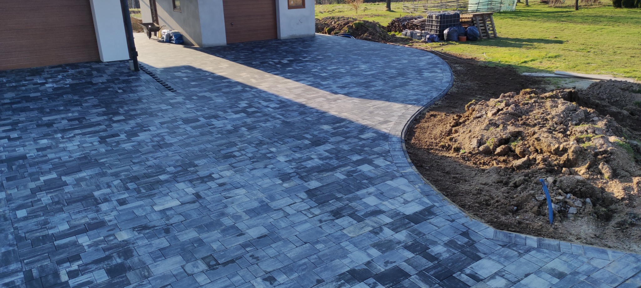 Niebiesko-szara kostka brukowa ułożona na podjeździe z widocznym odwodnieniem liniowym, obok świeżo usypana ziemia i trawnik.