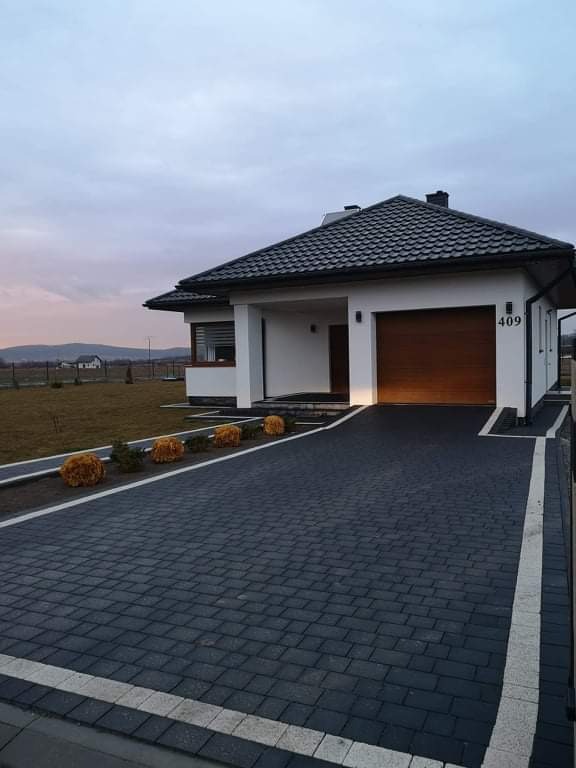 Nowoczesny dom z ciemnoszarą kostką brukową na podjeździe, obrzeża z jasnej kostki, oświetlony garaż, numer domu 409, widok na góry w tle.