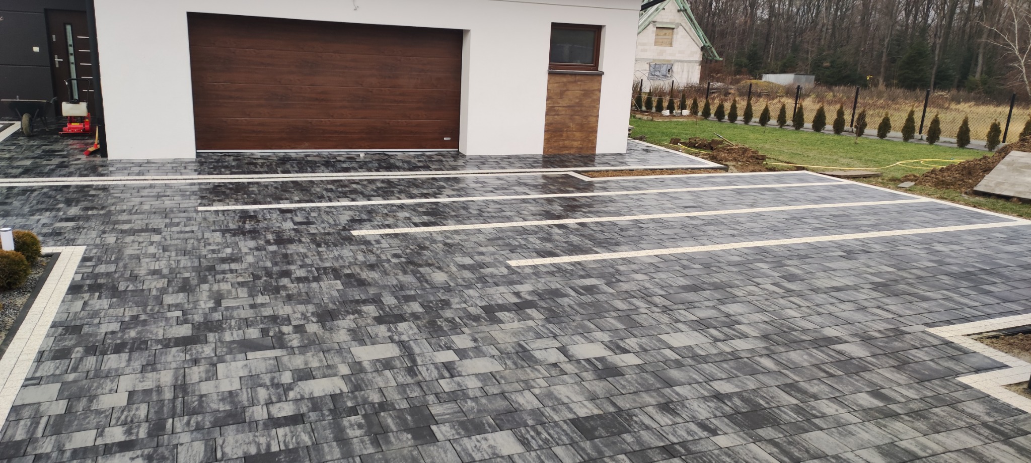 Ułożona kostka brukowa w odcieniach szarości i bieli na podjeździe przed domem z brązową bramą garażową, z widoczną w tle zielenią trawnika i drzew