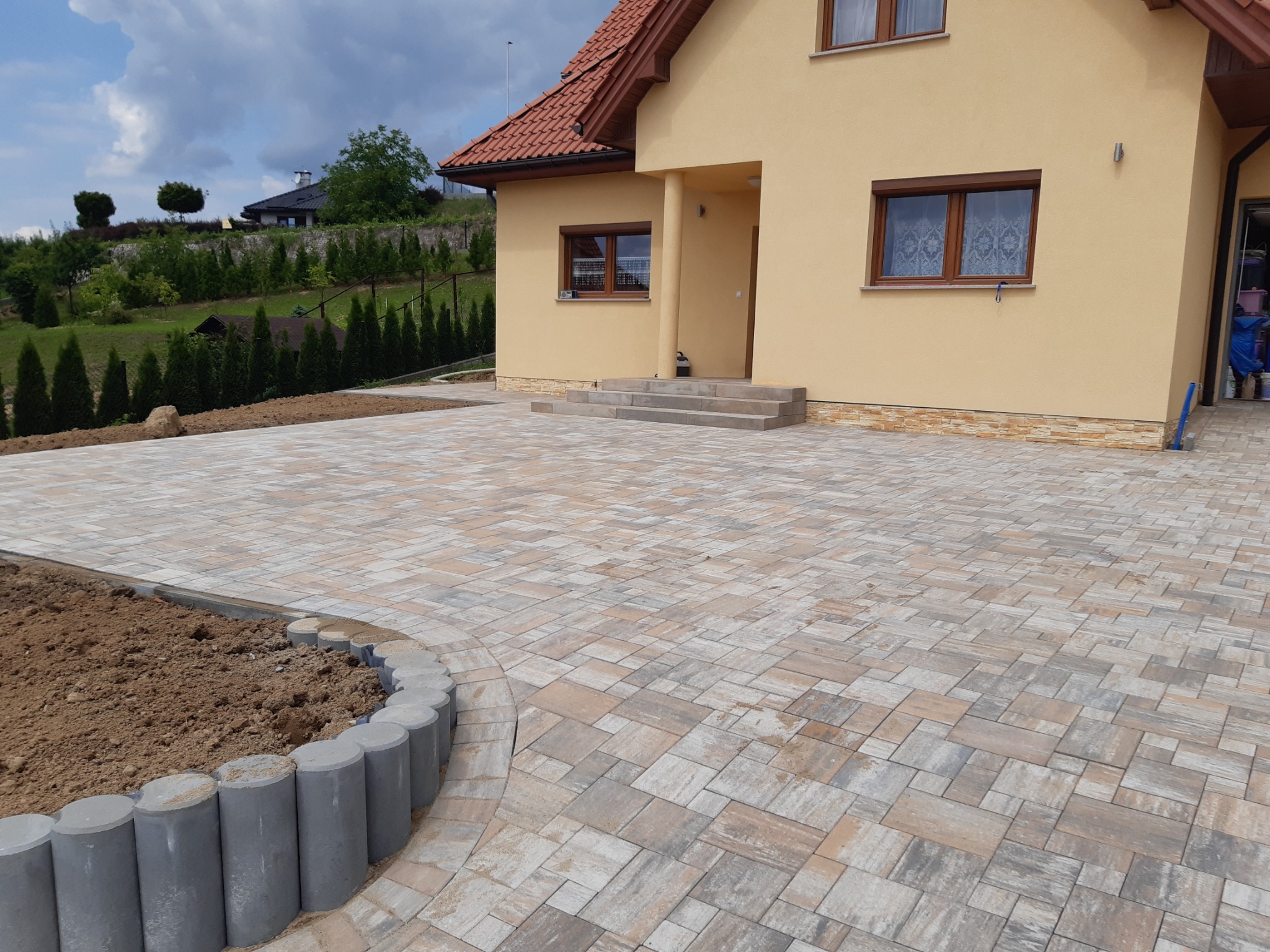 Nowo ułożona kostka brukowa w odcieniach szarości i beżu prowadząca do beżowego domu z brązowym dachem, obrzeże z betonowych palisad oddziela kostkę od rabaty z ziemią