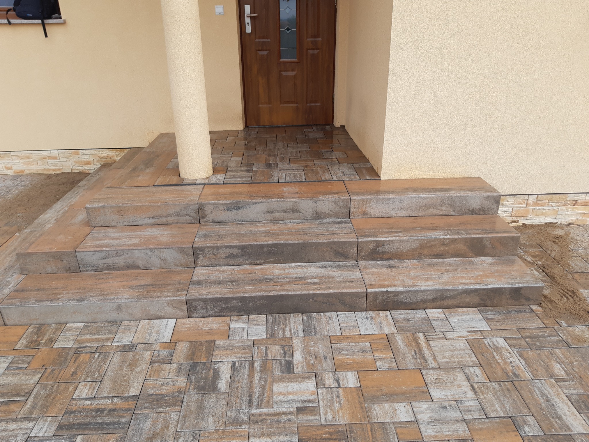 Schody wejściowe wyłożone kostką brukową w odcieniach brązu i szarości, prowadzące do drewnianych drzwi wejściowych budynku o beżowej elewacji.
