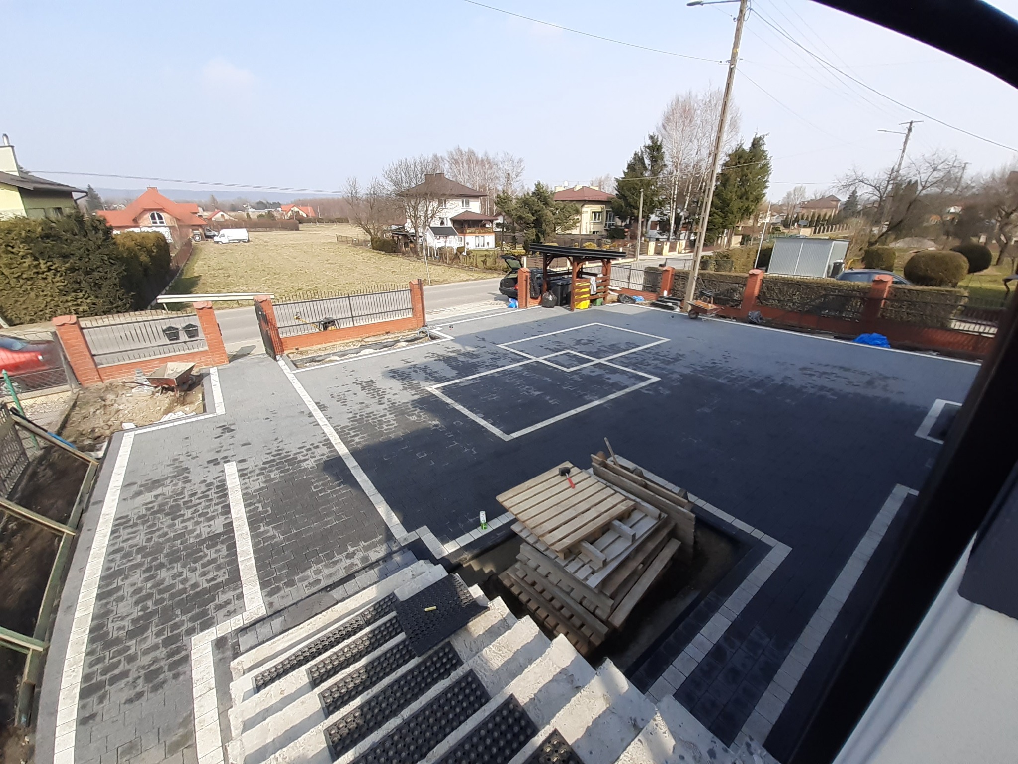 Nowa, ciemnoszara nawierzchnia z kostki brukowej z jasnymi obramowaniami, widok z góry na posesję z elementami krajobrazu w tle, palety z kostką opierają się o schody wejściowe.