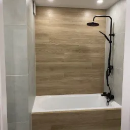 Nowoczesna łazienka z wanną obudowaną panelami drewnopodobnymi, czarna armatura i szare płytki, oświetlenie punktowe.