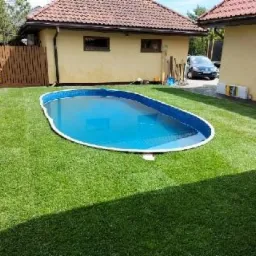 Owalny basen z błękitną wodą w ogrodzie z nowo założoną, intensywnie zieloną trawą z rolki, obok żółty budynek z brązowym dachem.