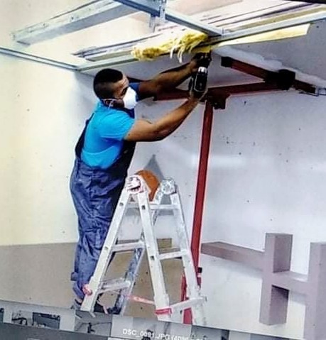 Mężczyzna w kombinezonie i masce ochronnej, stojący na aluminiowej drabinie, wierci w konstrukcji sufitu z widoczną izolacją z wełny mineralnej, używając wiertarki akumulatorowej.