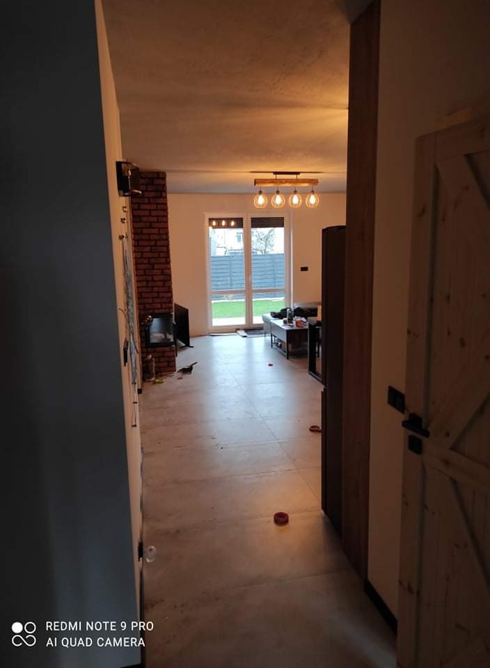 Wnętrze domu w trakcie wykańczania: widok na salon z kominkiem z cegły, nową podłogą z płytek, nowoczesną lampą wiszącą i częściowo otwartymi drzwiami z drewna w stylu rustykalnym.