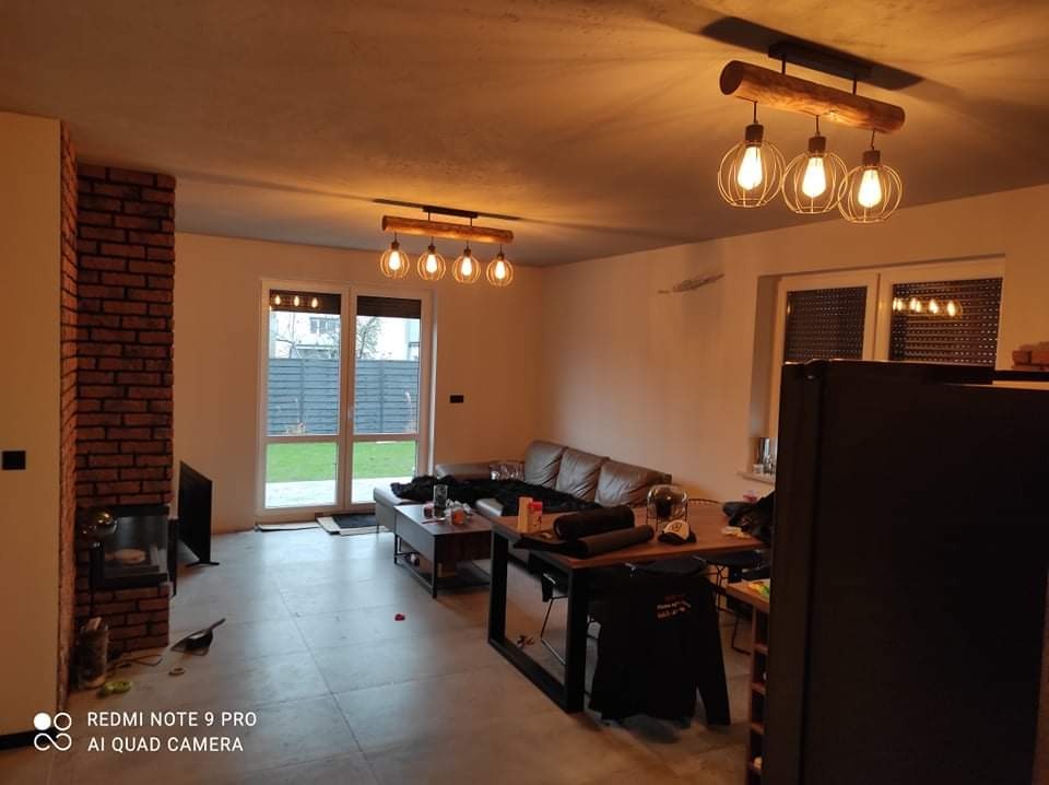 Salon po remoncie z ceglanym kominem, drewnianymi lampami wiszącymi, szarą podłogą i widokiem na ogród przez okno balkonowe.