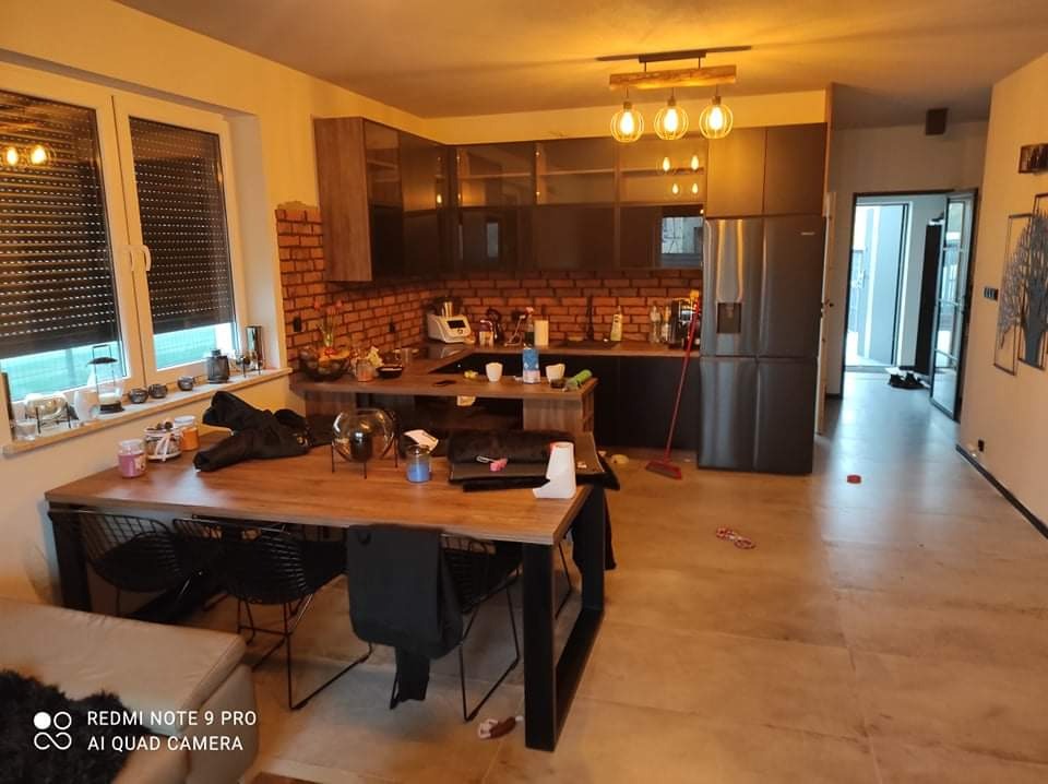 Nowoczesna kuchnia z ceglaną ścianą, drewnianym blatem i czarnymi szafkami, oświetlona wiszącymi lampami z żarówkami Edison, widok na jadalnię z drewnianym stołem i czarnymi krzesłami.