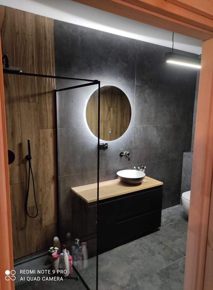 Nowoczesna łazienka z kabiną prysznicową typu walk-in, okrągłym lustrem z podświetleniem LED, umywalką nablatową na szafce z drewnianym blatem, szarą glazurą i drewnopodobnymi panelami ściennymi.