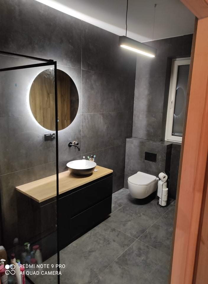 Nowoczesna łazienka z szarymi płytkami, okrągłym lustrem z podświetleniem LED, wiszącą toaletą i szafką pod umywalką z drewnianym blatem.