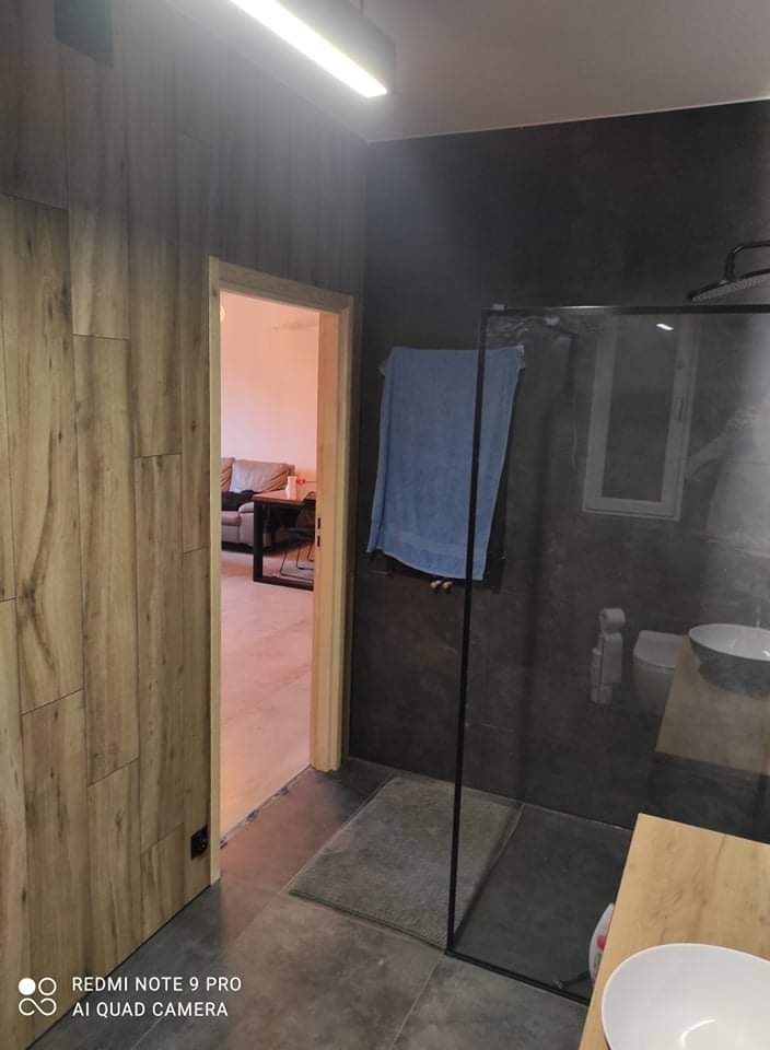 Nowoczesna łazienka z drewnopodobną okładziną ścienną, szarą kabiną prysznicową typu walk-in i widokiem na salon przez otwarte drzwi.