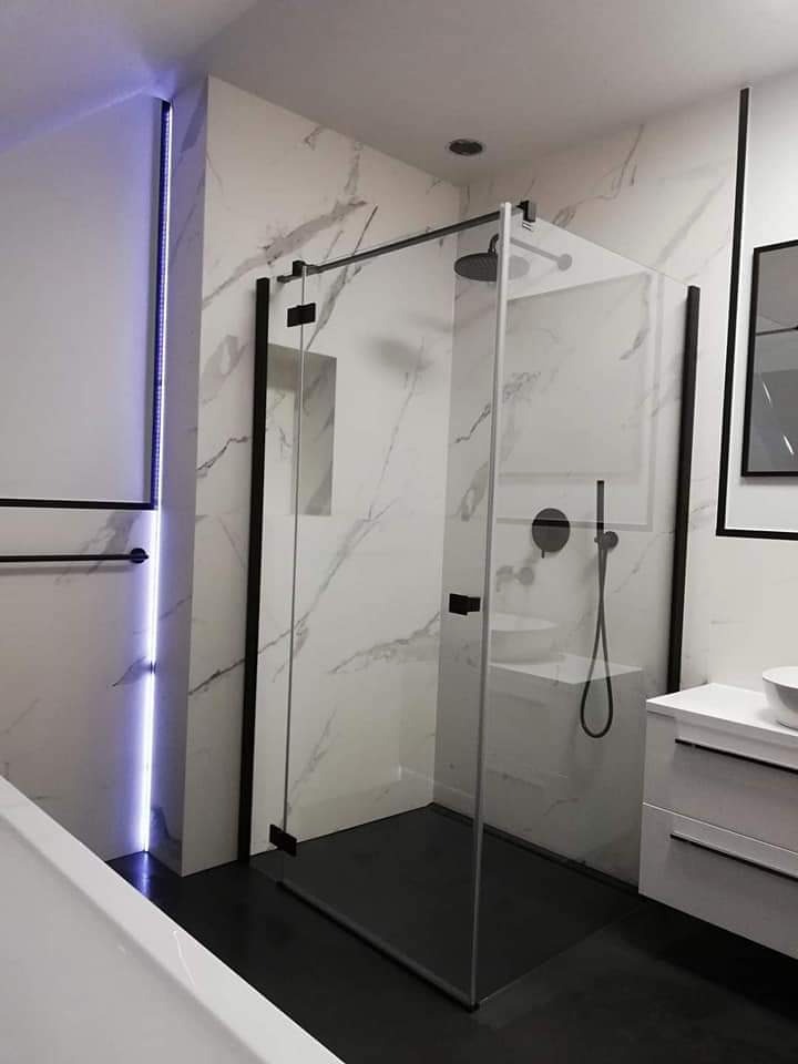 Nowoczesna kabina prysznicowa walk-in z czarnymi profilami na tle marmurowych płytek, podświetlenie LED w pionowej wnęce, czarna armatura i biała szafka umywalkowa.