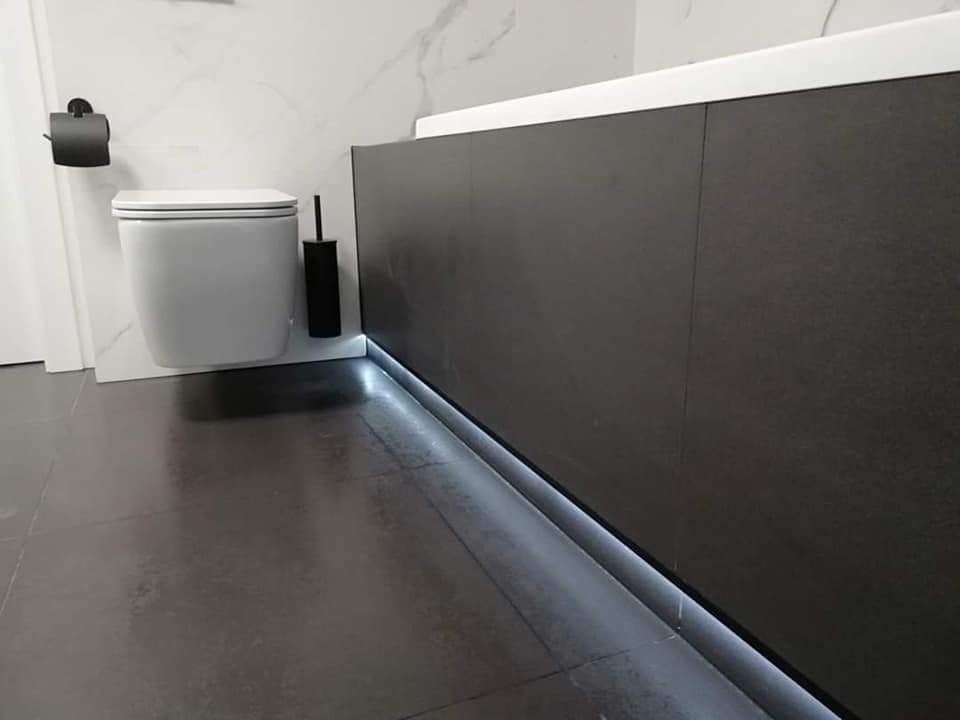 Nowoczesna łazienka z podświetleniem LED u dołu zabudowy wanny, wisząca toaleta z czarnym uchwytem na papier toaletowy i szczotką, ciemna, matowa podłoga.