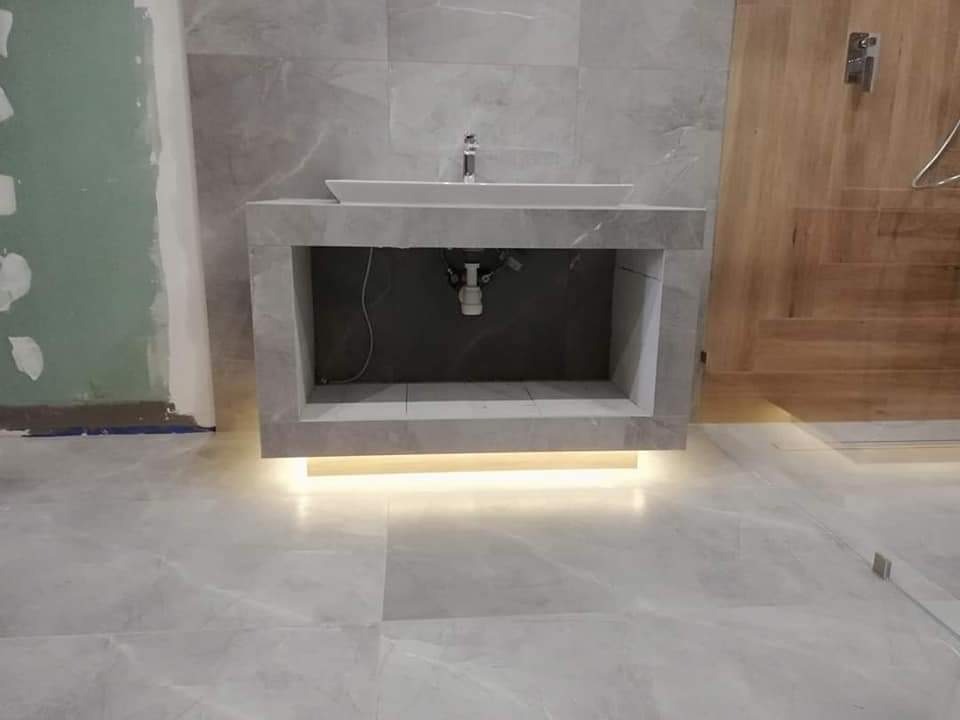 Łazienka w trakcie wykańczania: umywalka nablatowa na kamiennym blacie z podświetleniem LED, widoczne przyłącza hydrauliczne, ściany i podłoga wyłożone marmurowymi płytkami, obok drewniane drzwi...