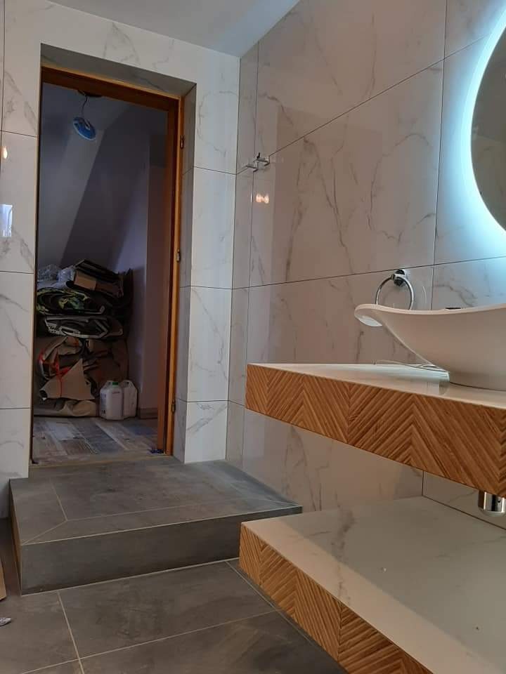 Wykończona łazienka we Wrześni z marmurowymi płytkami, drewnianymi akcentami na szafkach umywalkowych i widocznym wejściem do poddasza z materiałami budowlanymi.