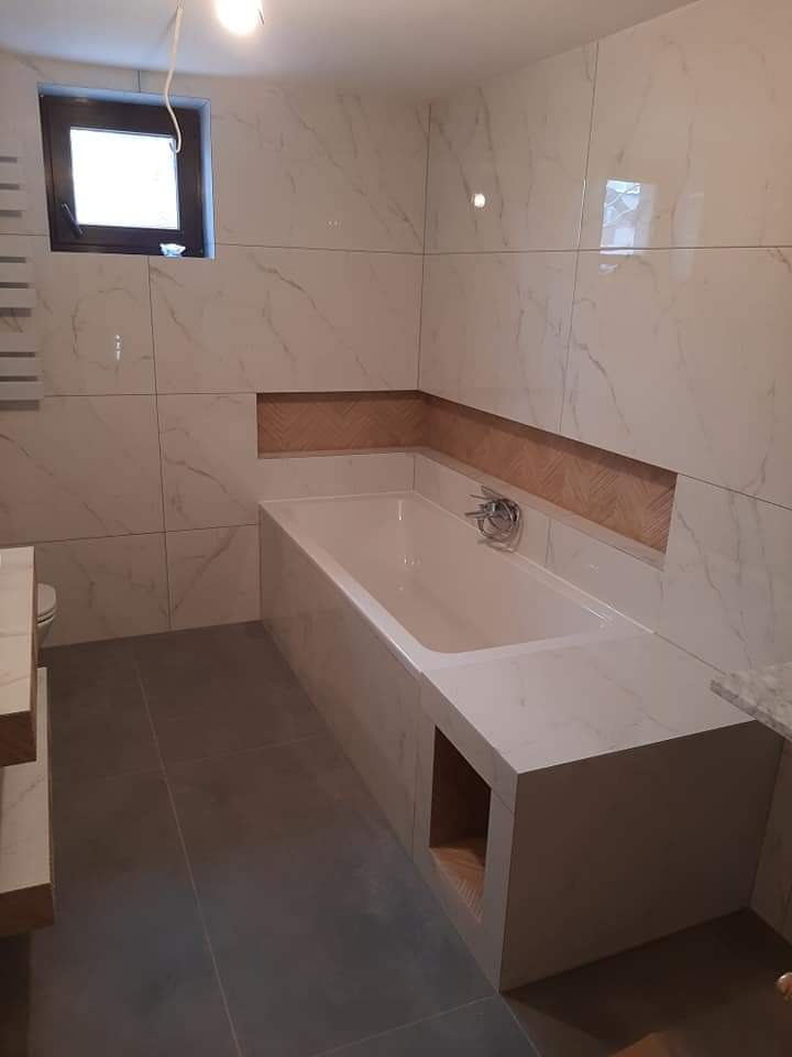 Nowoczesna łazienka z wanną obudowaną płytkami imitującymi marmur, szare płytki podłogowe, małe okno i wnęka nad wanną z dekoracyjną okładziną.