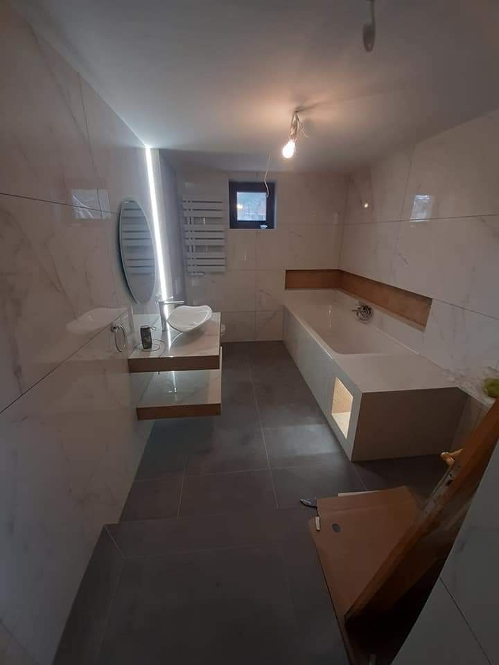 Nowoczesna łazienka z białymi marmurowymi płytkami, szarą podłogą, owalnym lustrem i minimalistyczną umywalką, widoczne surowe wykończenie wanny z podświetleniem, wisząca żarówka i małe okno.