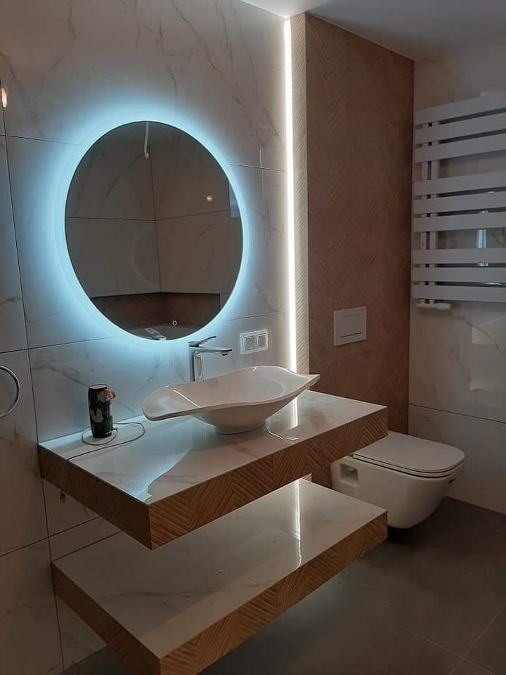 Nowoczesna łazienka z okrągłym lustrem podświetlanym LED, umywalką w kształcie liścia na blacie imitującym marmur i drewno, oraz wiszącą toaletą.