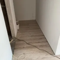 Wnętrze w trakcie remontu: ułożone panele podłogowe w odcieniach brązu, białe ściany częściowo pomalowane, widoczne przewody elektryczne i wiadro z narzędziami.