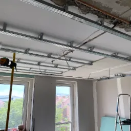 Wnętrze w trakcie remontu: widok na metalowy stelaż sufitu podwieszanego z płyt kartonowo-gipsowych, laser poziomujący na statywie przy oknie, ściany częściowo obłożone płytami, drabina z narzędziami.