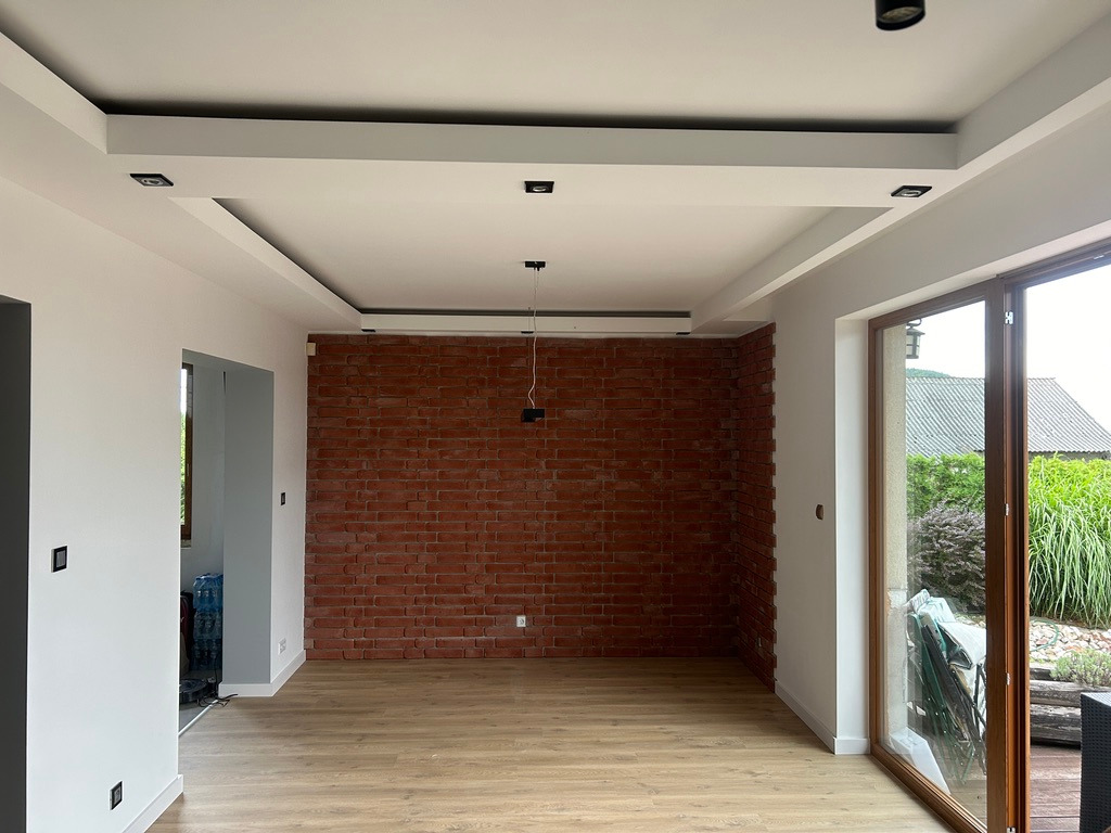 Wykończone wnętrze: parkiet, ceglana ściana, białe ściany i sufit z oświetleniem punktowym. Duże okno z widokiem na zieleń. Minimalistyczny design.