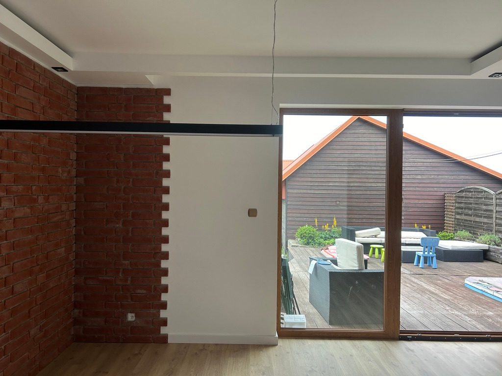 Ceglana ściana z białym fragmentem, widok na taras przez duże okno. Elementy dekoracyjne i drewniana podłoga tworzą nowoczesne wnętrze.