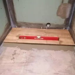 Przygotowanie podłoża pod prysznic: ułożone płytki imitujące drewno, poziomica czerwona na wierzchu, widoczne krzyżyki dystansowe, surowa ściana w tle.