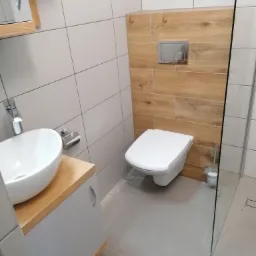 Nowoczesna łazienka z umywalką nablatową, podwieszaną toaletą i płytkami imitującymi drewno na ścianie za sedesem, szara podłoga i kabina prysznicowa ze szkła.