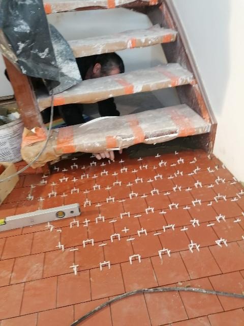 Montaż terakoty na podłodze pod schodami, widoczny poziomica i krzyżyki dystansowe, glazurnik w trakcie pracy.