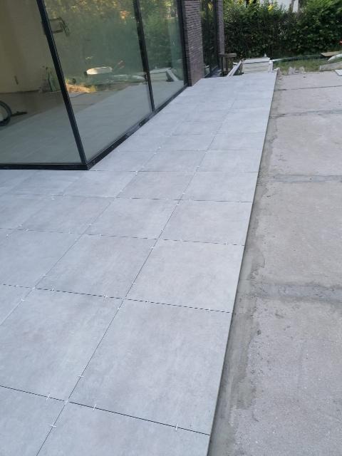 Ułożone szare płytki na zewnątrz budynku, z widocznymi krzyżykami dystansowymi, przylegające do betonowej powierzchni i przeszklenia.