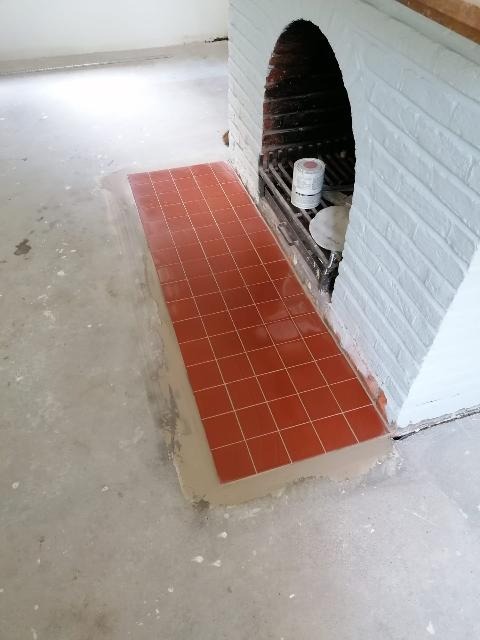 Pomarańczowe płytki podłogowe ułożone przed kominkiem z białej cegły, widoczne ślady zaprawy, etap prac wykończeniowych.