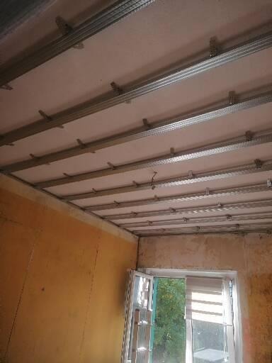 Montaż metalowej konstrukcji sufitu podwieszanego z płyt kartonowo-gipsowych w pomieszczeniu z oknem, widoczne profile i uchwyty mocujące do stropu.