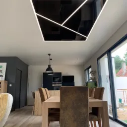 Nowoczesny salon z jadalnią, na suficie podwieszany, czarny element z geometrycznym oświetleniem LED, widok na kuchnię i duże okno tarasowe.