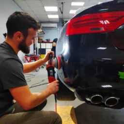 Detailer poleruje czarny lakier samochodu przy użyciu czerwonej polerki rotacyjnej, widoczne odbicia światła na karoserii i podwójny wydech.