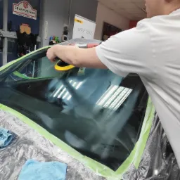 Polerowanie przedniej szyby samochodu w warsztacie z użyciem polerki elektrycznej Mirka Deros; szyba zabezpieczona taśmą malarską, widoczne mikrofibry i folia ochronna.