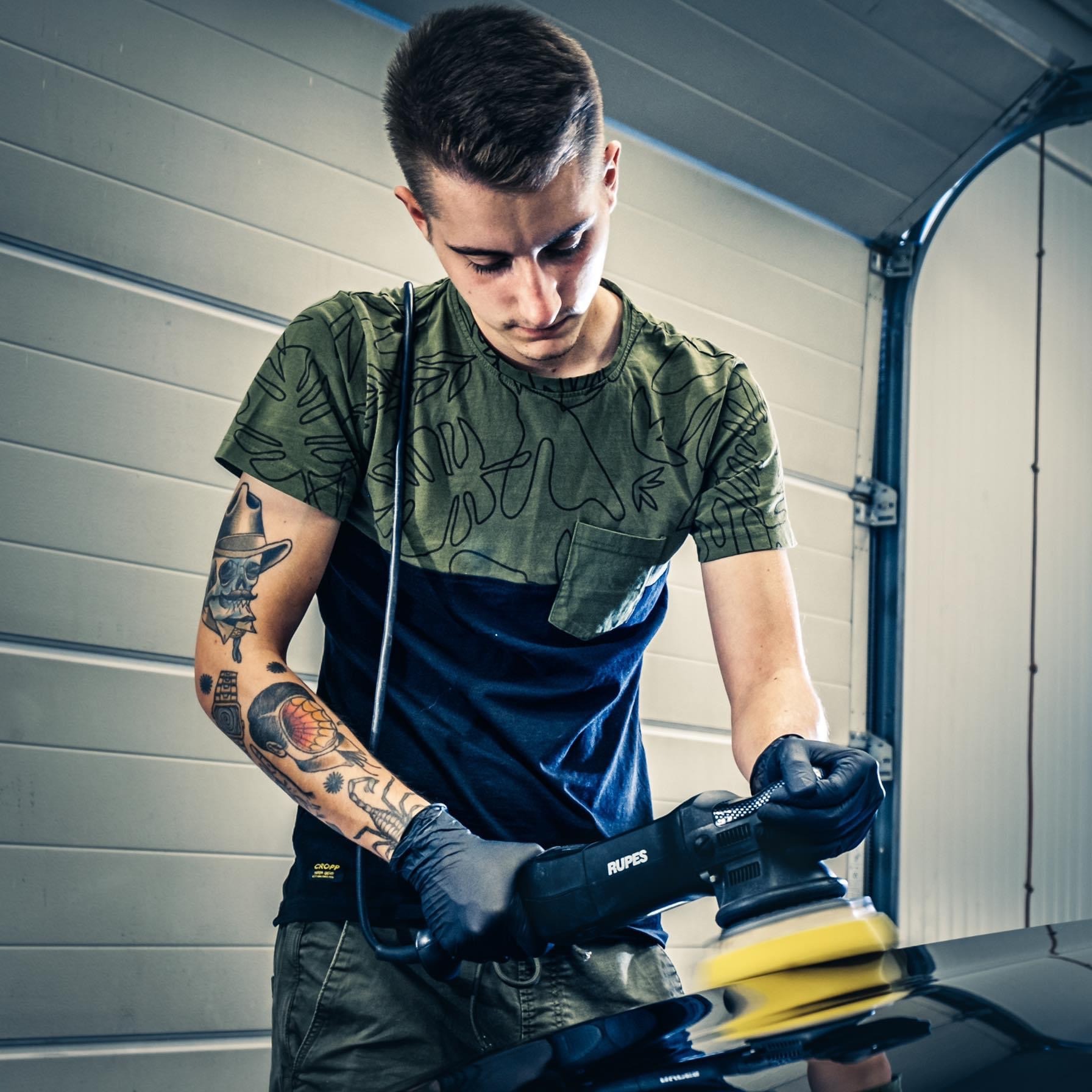 Młody mężczyzna z tatuażami na ramieniu poleruje czarny lakier samochodu profesjonalną polerką Rupes w garażu.