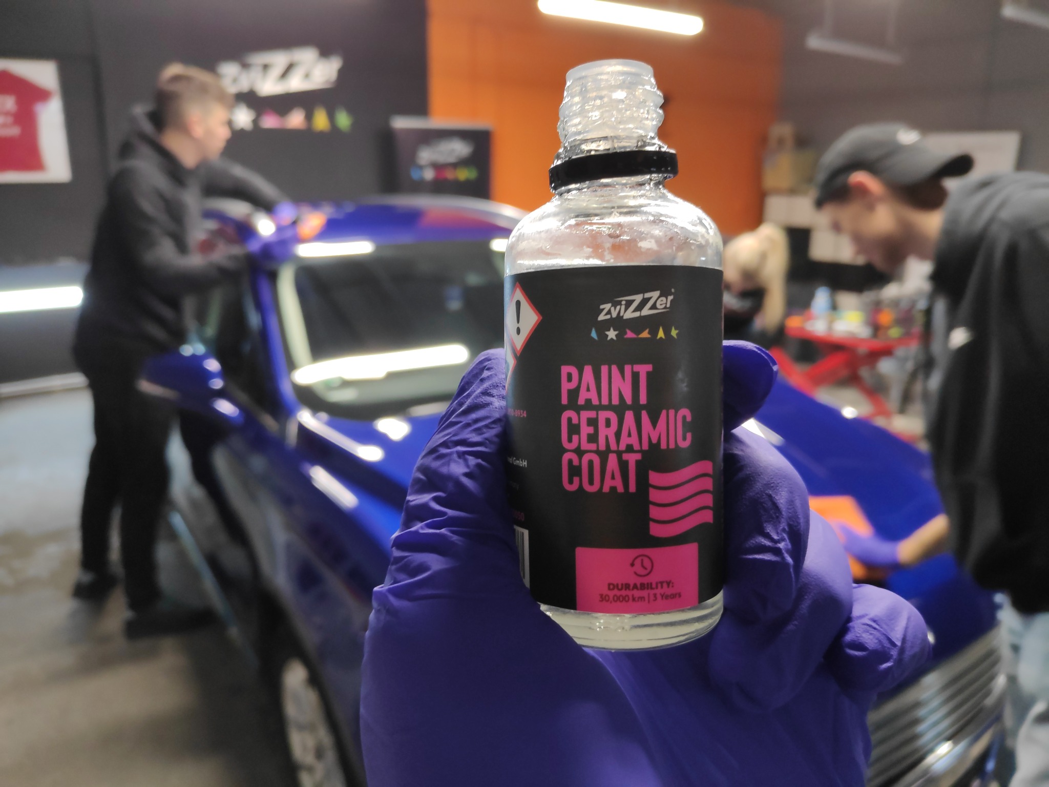 Aplikacja ceramicznej powłoki ochronnej ZviZZer na niebieski lakier samochodu, widoczna butelka produktu trzymana w dłoni w fioletowej rękawicy, w tle pracownicy polerujący auto.