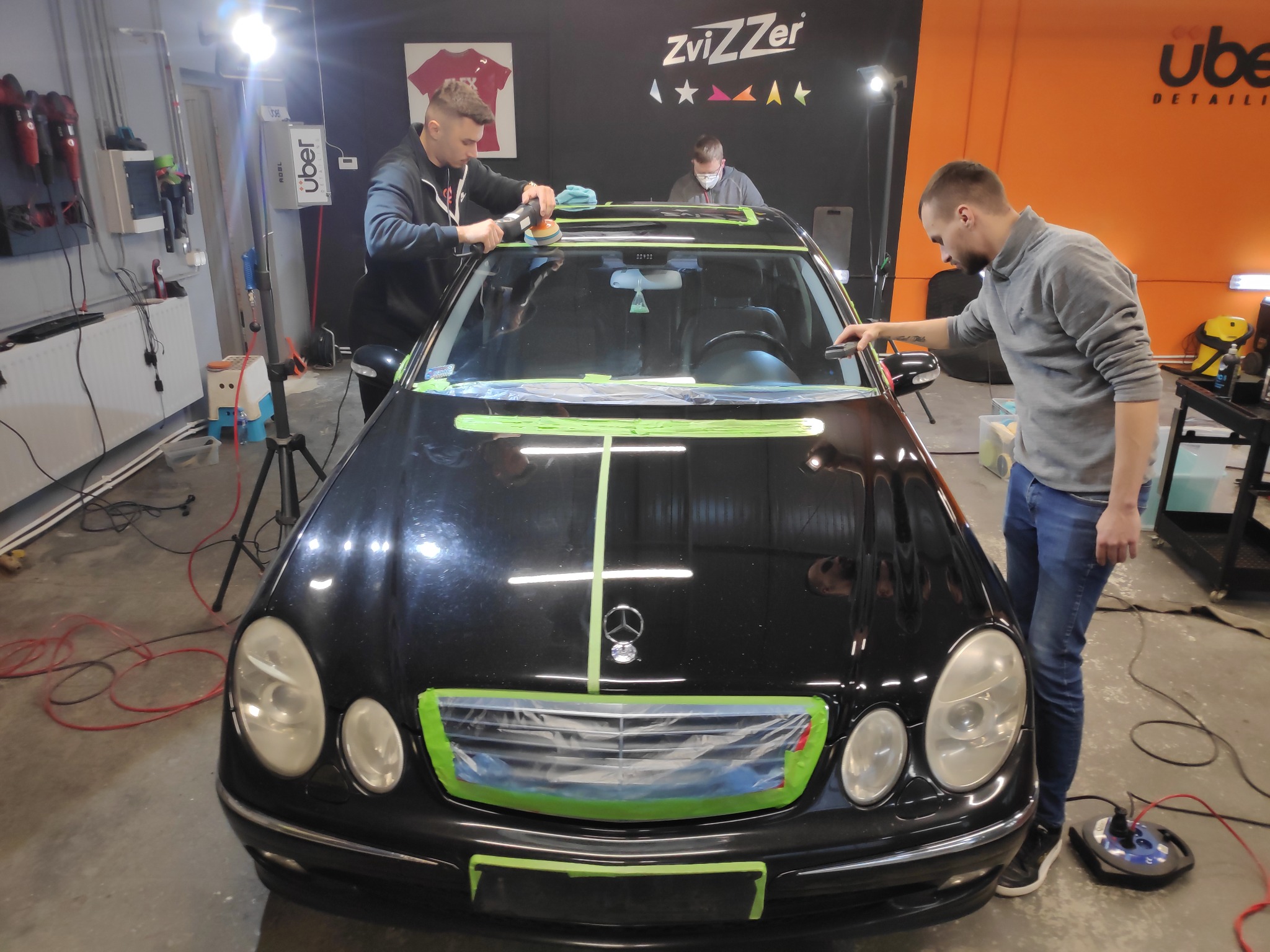 Polerowanie karoserii czarnego Mercedesa E-klasy w warsztacie detailingu, z użyciem taśmy maskującej i polerki rotacyjnej, widoczne logo Zvizzer na ścianie.