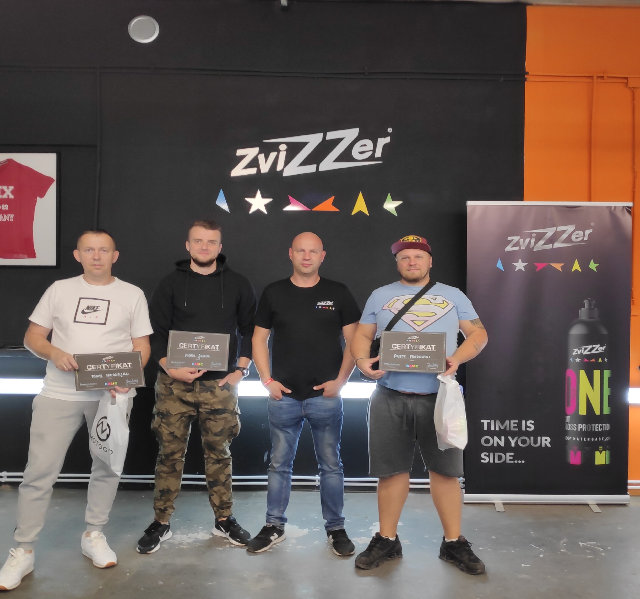 Czterech mężczyzn pozuje z certyfikatami firmy ZviZZer na tle czarno-pomarańczowej ściany z logo firmy i banerem reklamowym produktu ONE.