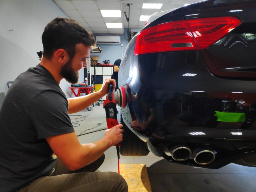 Detailer poleruje czarny lakier samochodu przy użyciu czerwonej polerki rotacyjnej, widoczne odbicia światła na karoserii i podwójny wydech.