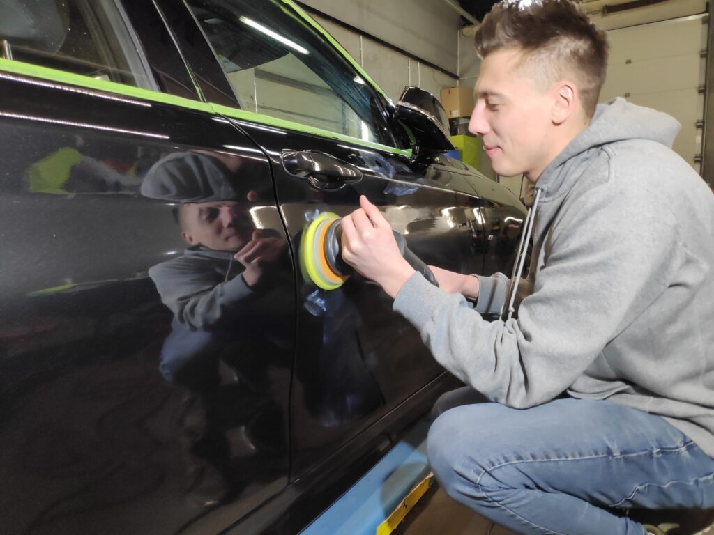 Młody mężczyzna poleruje czarny lakier samochodu przy użyciu polerki rotacyjnej, widoczne odbicie jego sylwetki na karoserii.
