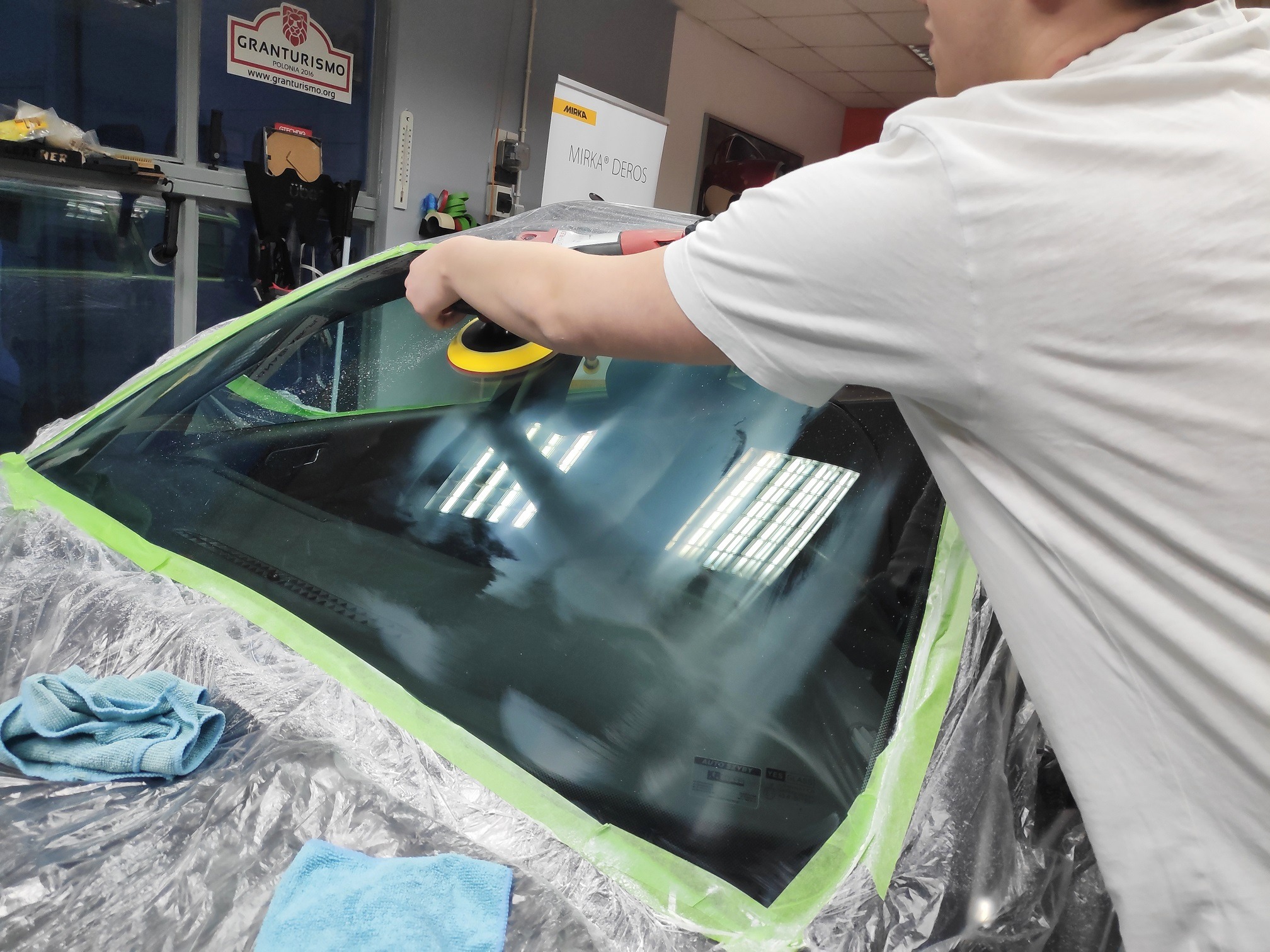 Polerowanie przedniej szyby samochodu w warsztacie z użyciem polerki elektrycznej Mirka Deros; szyba zabezpieczona taśmą malarską, widoczne mikrofibry i folia ochronna.