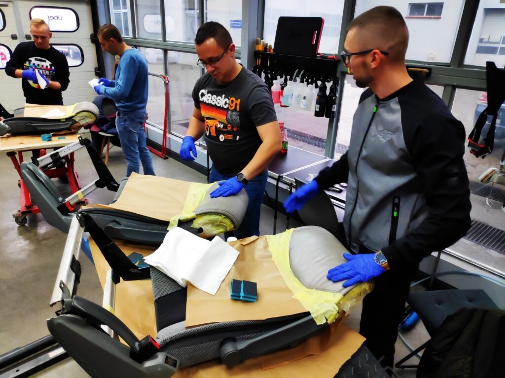 Trzech mężczyzn w niebieskich rękawiczkach pracuje nad renowacją samochodowych siedzeń w warsztacie; widoczne narzędzia i materiały zabezpieczające.