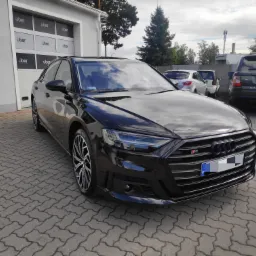 Błyszczący, czarny sedan Audi S8 zaparkowany przed myjnią Uber Detailing, z widocznym odbiciem chmur w karoserii i kostką brukową pod kołami.