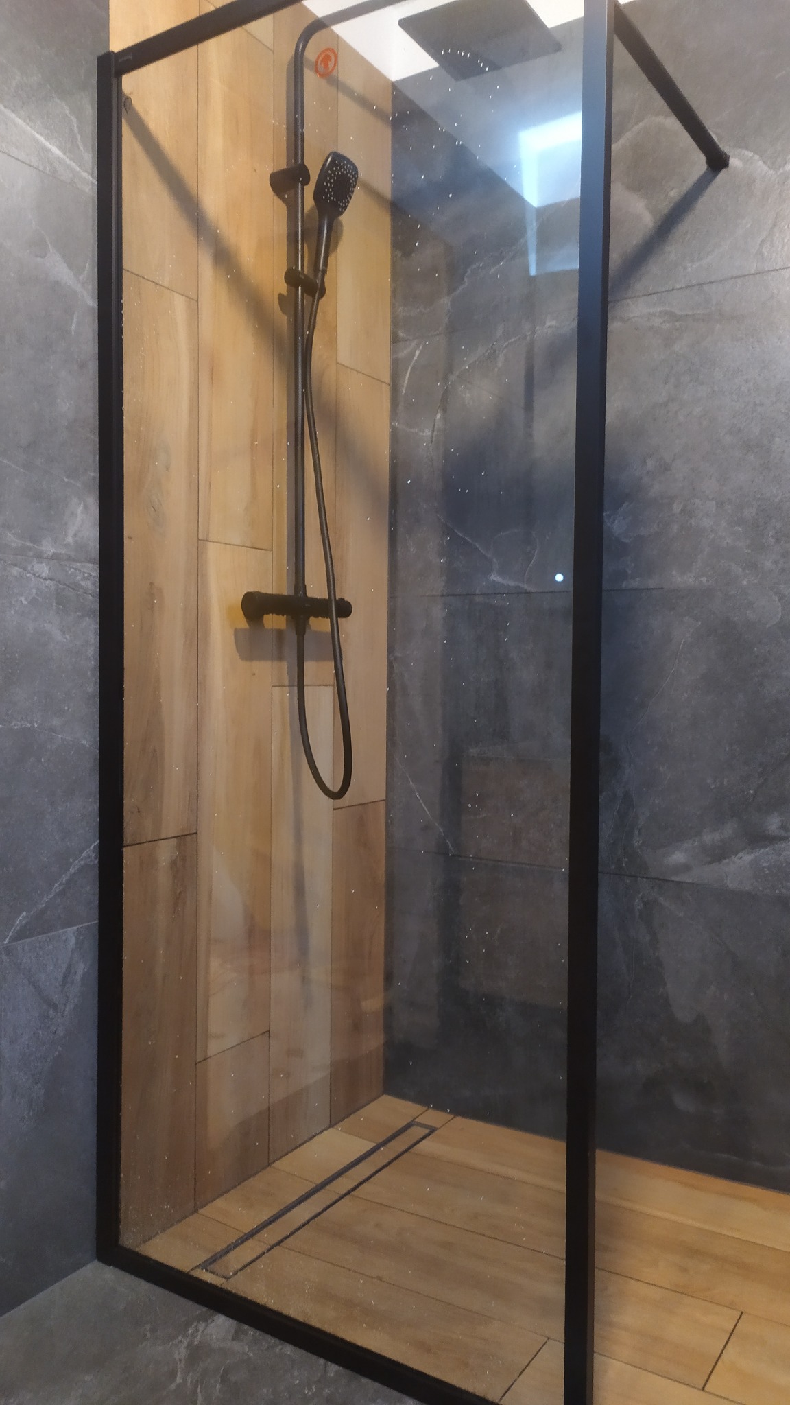 Nowoczesna kabina prysznicowa z czarną armaturą i drewnopodobnymi płytkami na ścianach i podłodze, oddzielona szybą w czarnej ramie, na tle szarych płytek imitujących kamień.