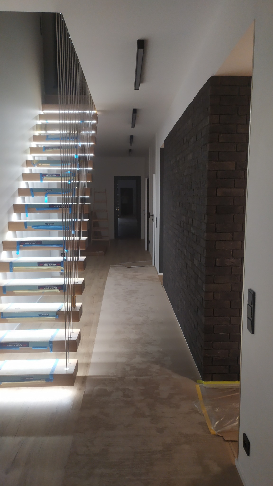 Korytarz w trakcie wykańczania: podłoga zabezpieczona folią, schody z podświetleniem LED zabezpieczone taśmą malarską, ściana wyłożona cegłą klinkierową w odcieniach brązu i grafitu.